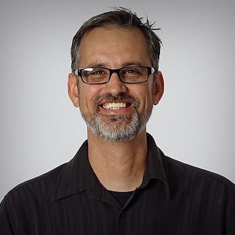 A smiling person wearing glasses and a dark shirt.