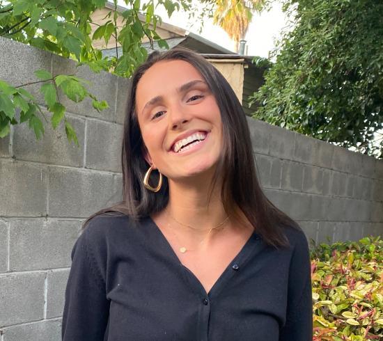 A person smiling in front of a stone wall and greenery.