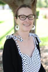 Headshot of smiling woman with a light complexion and light brown hair pulled back with a black hair clip. She is wearing cat eye glasses, a white dress with black polka dots, a black cardigan sweater, and dangling white circular earrings. There is an out-of-focus tree in the background.