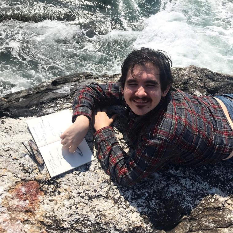 A person lying on rocks near water, holding a notebook.