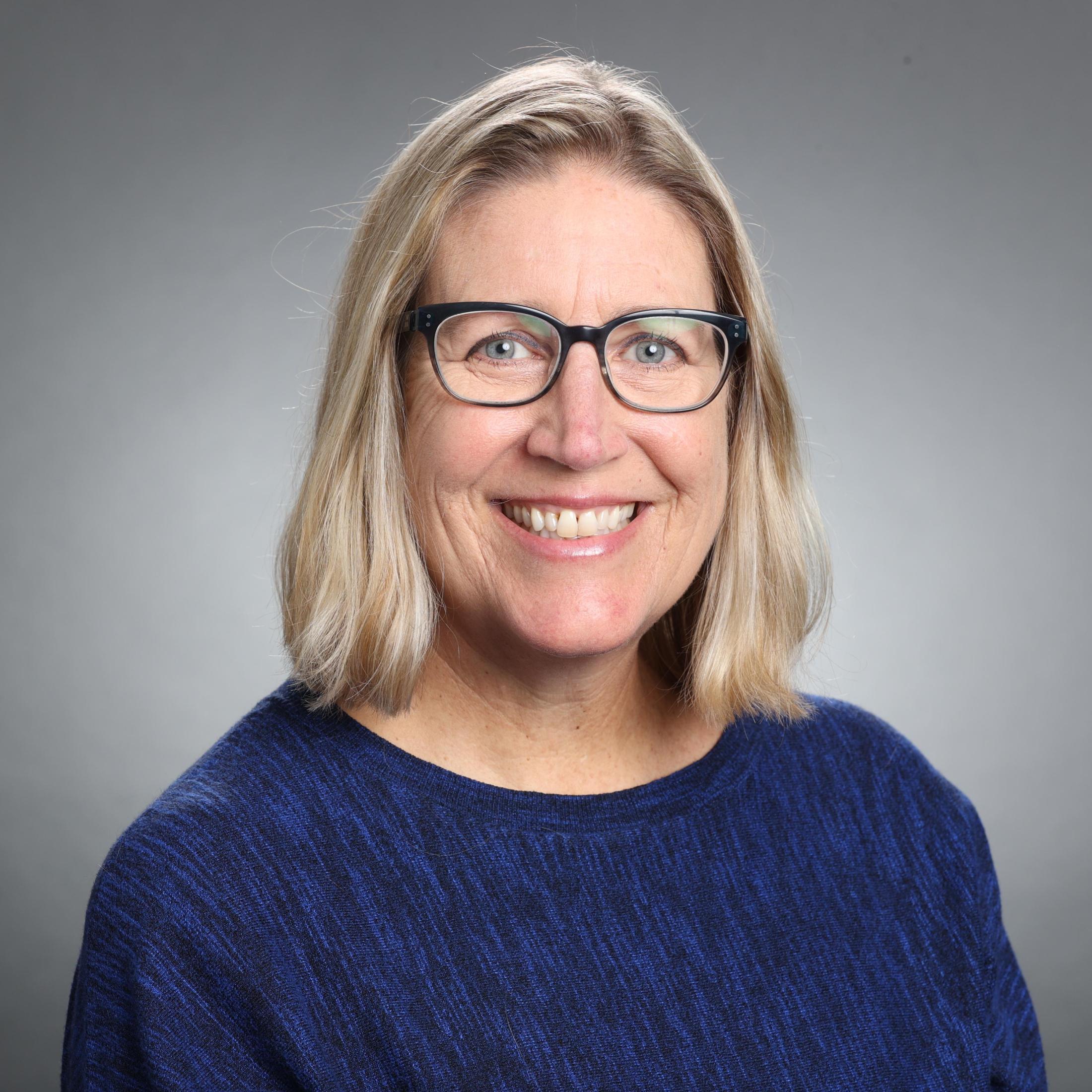 A person smiling, wearing glasses and a blue shirt against a grey background.