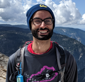 A person wearing glasses and a beanie, smiling outdoors with mountains in the background.