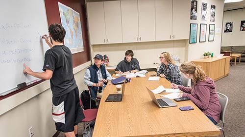Several students studying at a large table; one writes on a whiteboard. 
