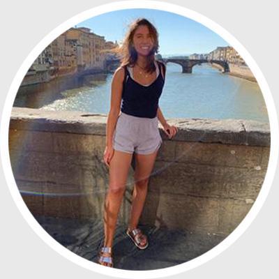 A woman stands by a river with a bridge in the background.