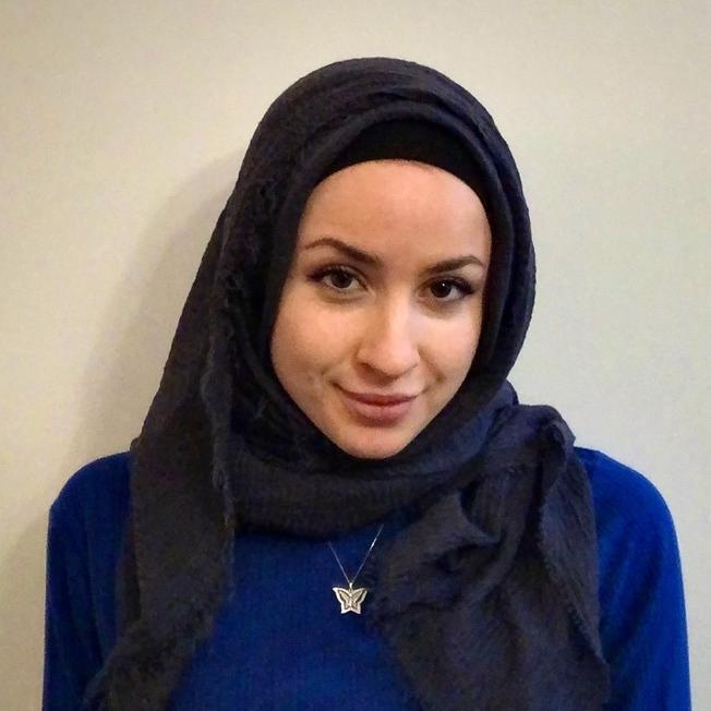 A woman in a blue top and dark headscarf against a plain wall.
