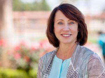 A woman smiling outdoors with flowers and greenery in the background.