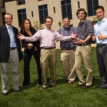 Group of six people standing on grass outside a building. 