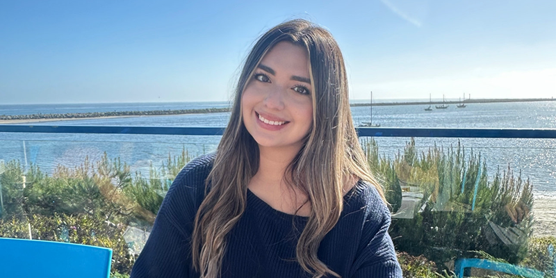 Alexis sits at an outdoor dining table with the water and sky in the background 