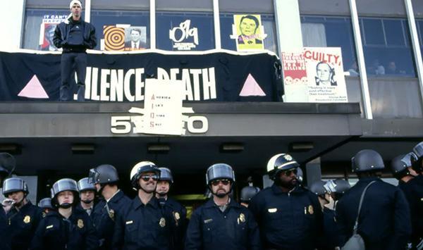 Police officers standing beneath balcony with protest signs reading 'DEPLOY FDA' and 'S.O.S.'