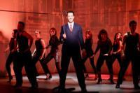 A scene from American Psycho with a man in a grey pinstripe suit and red tie flanked by an ensemble of male and female dancers in black