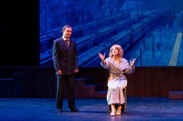 A man in a suit standing on the left and a woman in in a trench coat  gesticulating and seated on the right against a bright blue background