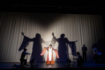 A woman in a white dress with a red robe over it trimmed with feathers in front of a white curtain projecting dancing shadows