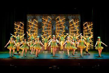 Male and female tap dancers wearing gold and green costumes in the big  42nd Street dance number set against a backdrop of lightbulbs in fan shapes