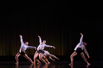 4 female dancers in white shirts and black shorts on a black stage
