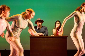 2 female dancers in white unitards dance in front of a table with a seated man in a grey suit and hat and a woman in a red dress