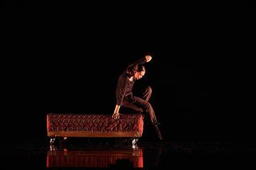 A female dancer in black tshirt and leggings sitting on the end of a red upholstered bench with casters