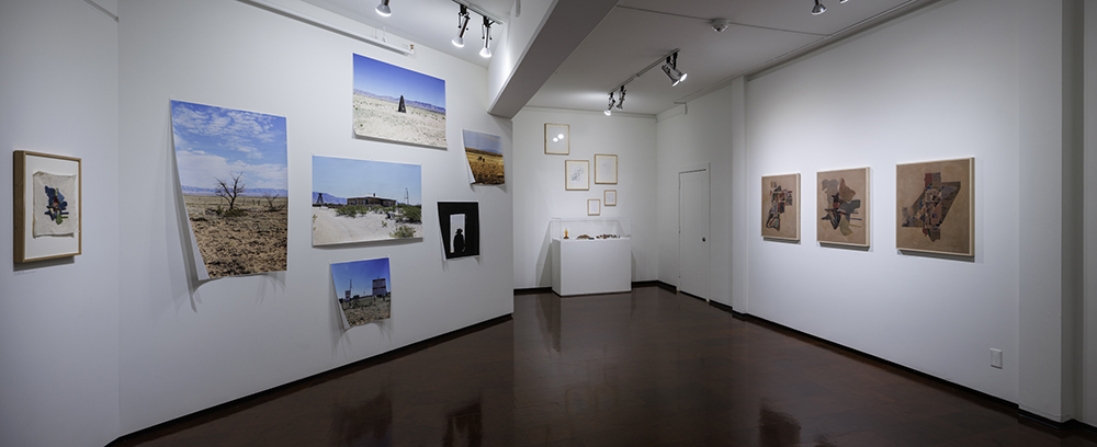 Gallery installation with photographs tacked to wall, framed artworks, and a display case