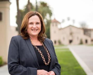 A woman in professional attire smiling outdoors near a building.