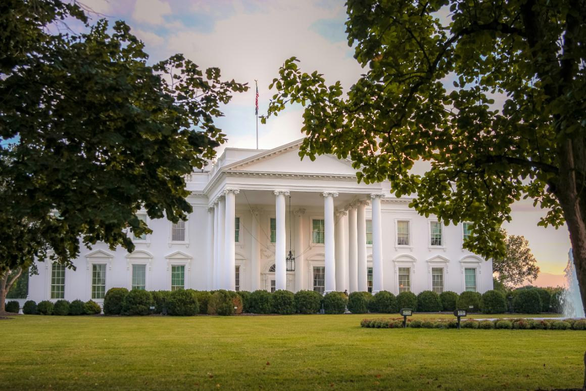 A large white house with columns and many windows. The house sits on a expansive green lawn with a large circular fountain and is framed by two green leafy trees.