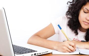 Student writing on paper next to a laptop.