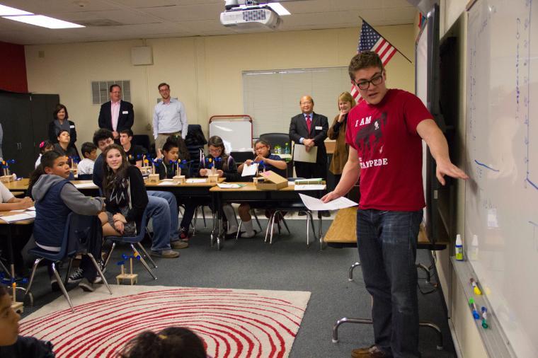 A teacher stands at the front of a classroom with students.