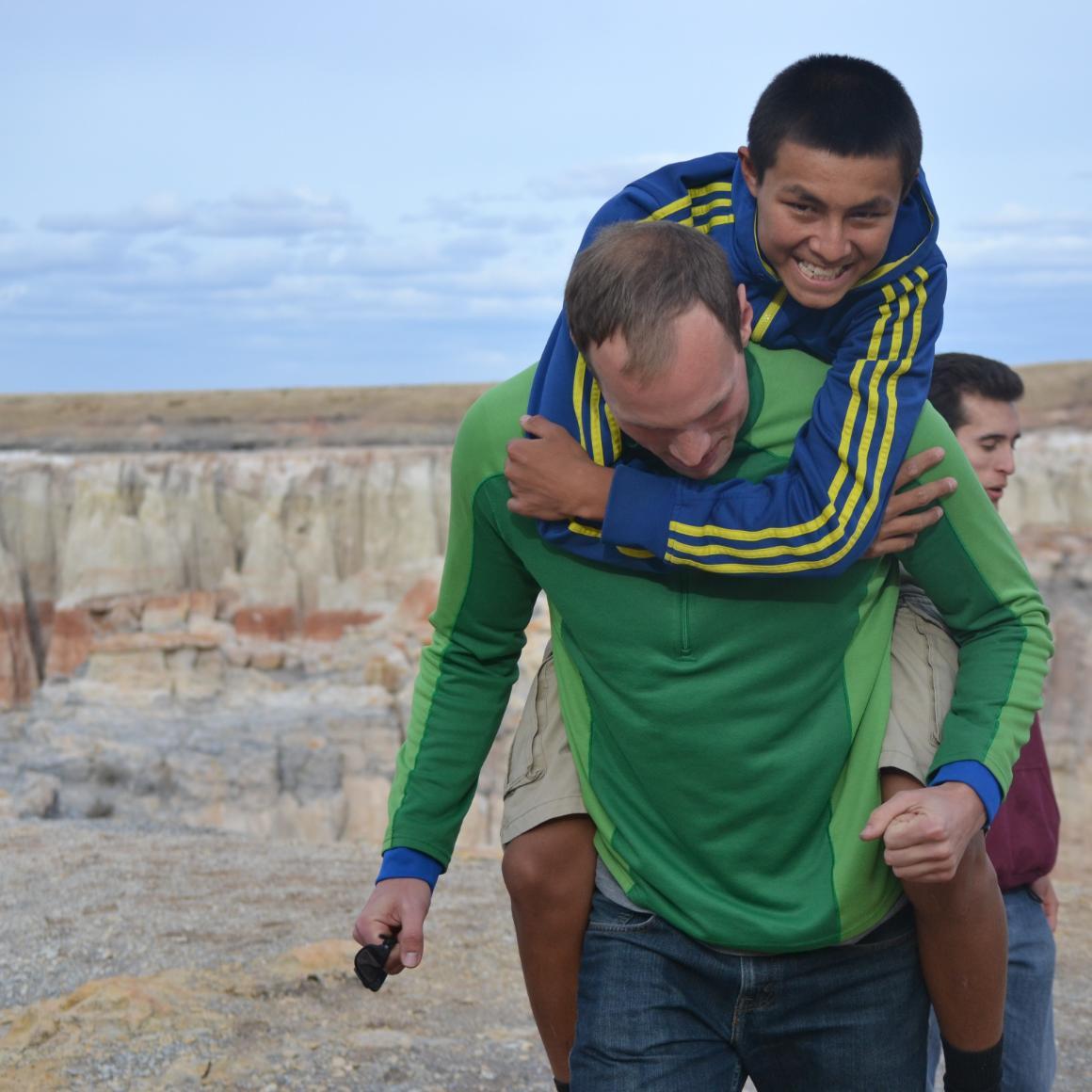 Two people, one carrying another on their back, near cliffs and the sea. 