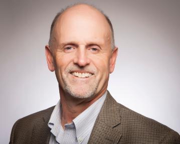 A person smiling, wearing a blazer and shirt, with a plain background.
