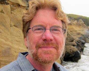 A person with glasses and a beard standing near rocky shore.