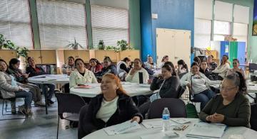 Camino Al Bienestar Group of community members in large room round tables.