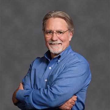 A man in a blue shirt smiling with arms crossed.