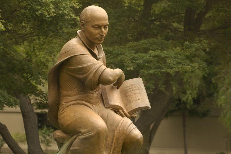 Statue of a Jesuit holding a book, surrounded by trees.