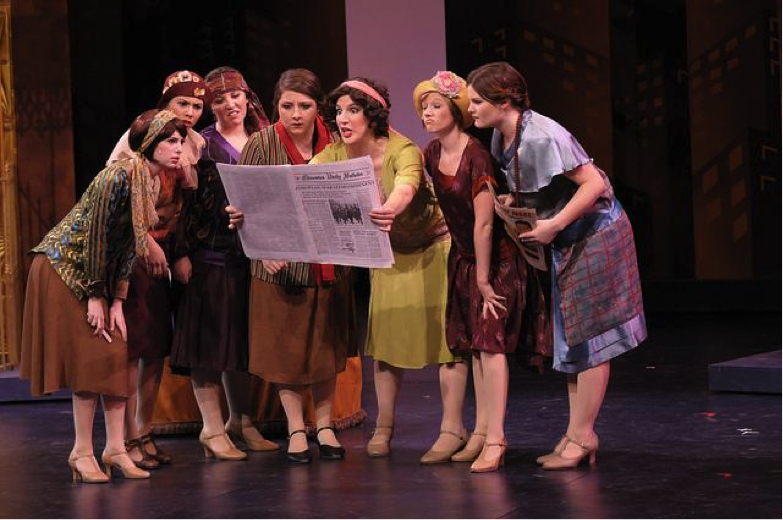 Several performers in period costumes gather to read a large paper on stage.