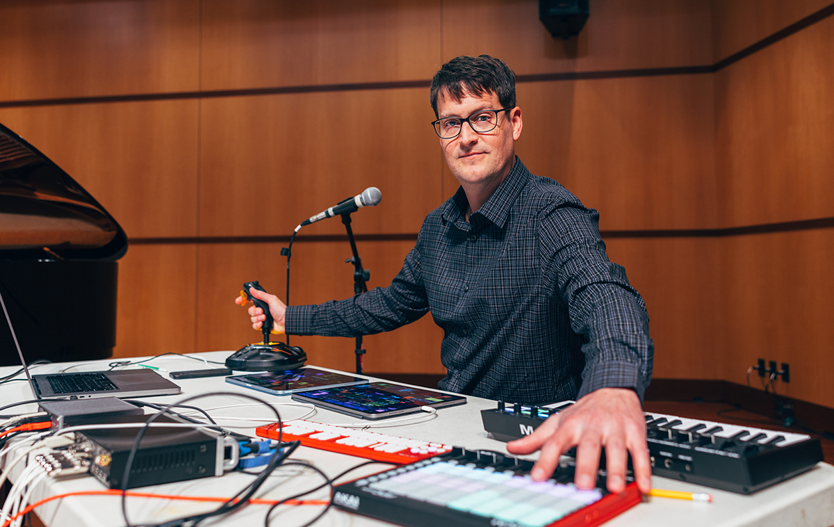 Sam Pluta interacts with microphone, joystick, keyboard and other electronic musical devices on a table in a performance hall.