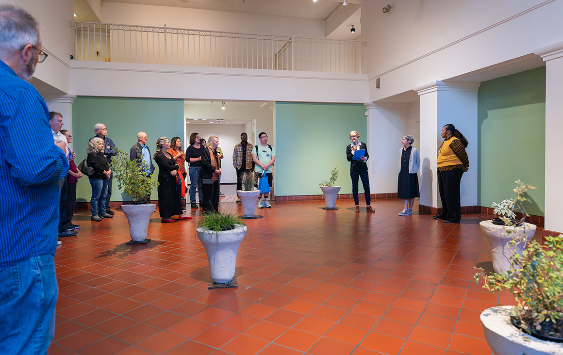 A group of guests stand in a semi-circle in the de Saisset Museum lobby, with bell-shaped adobe planters arranged across the middle of the lobby space.