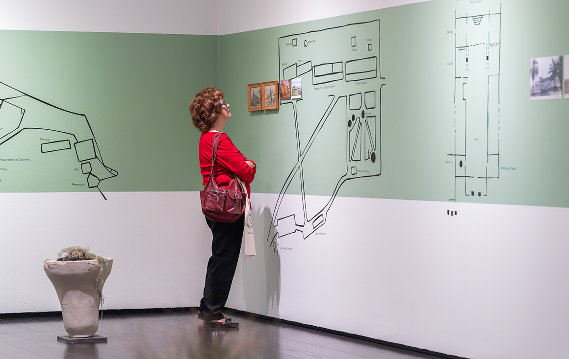 An older woman examines several aerial spec maps of Mission structures painted on the green gallery wall.