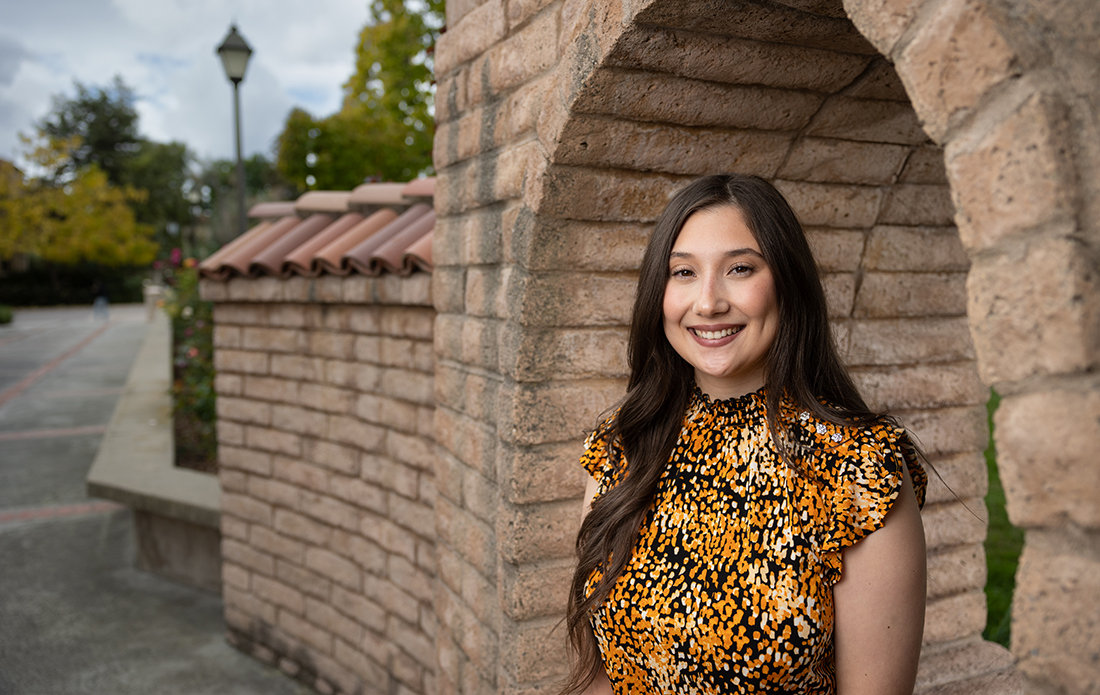 Leandra Couto sitting on brick wall on Santa Clara campus with sky in background