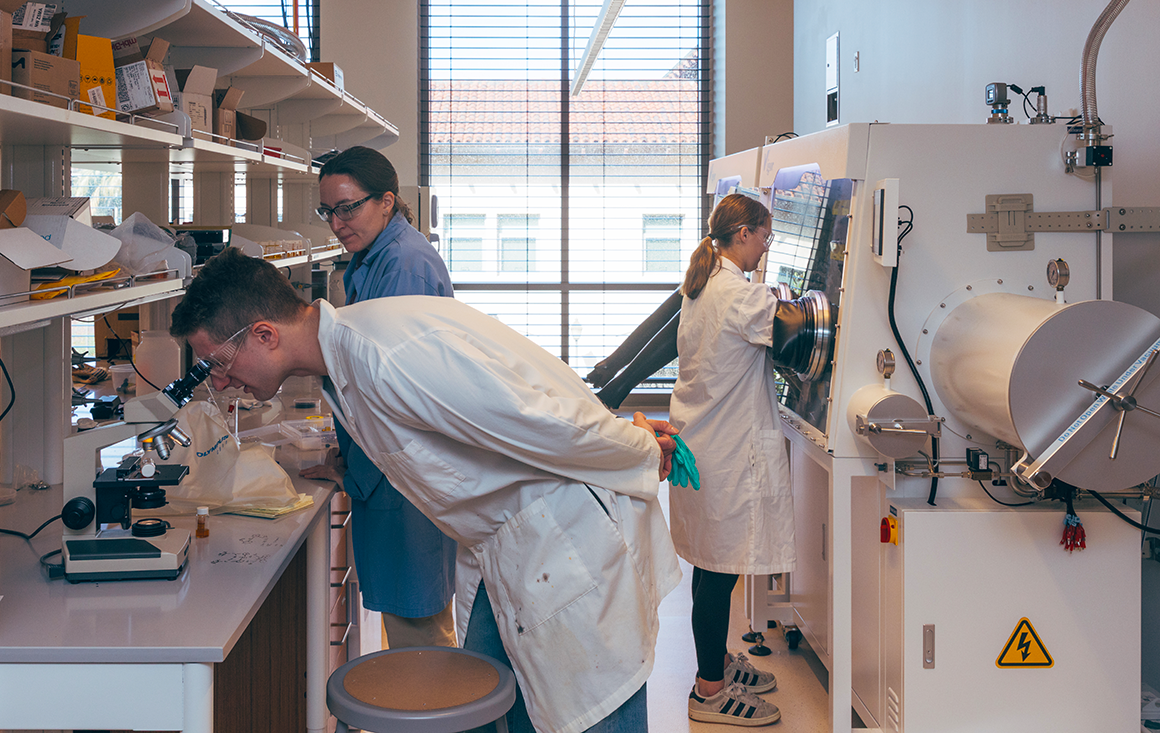 A wide shot of a chemistry lab with a professor supervising two students—one hovering over a microscope, the other working in a glovebox.