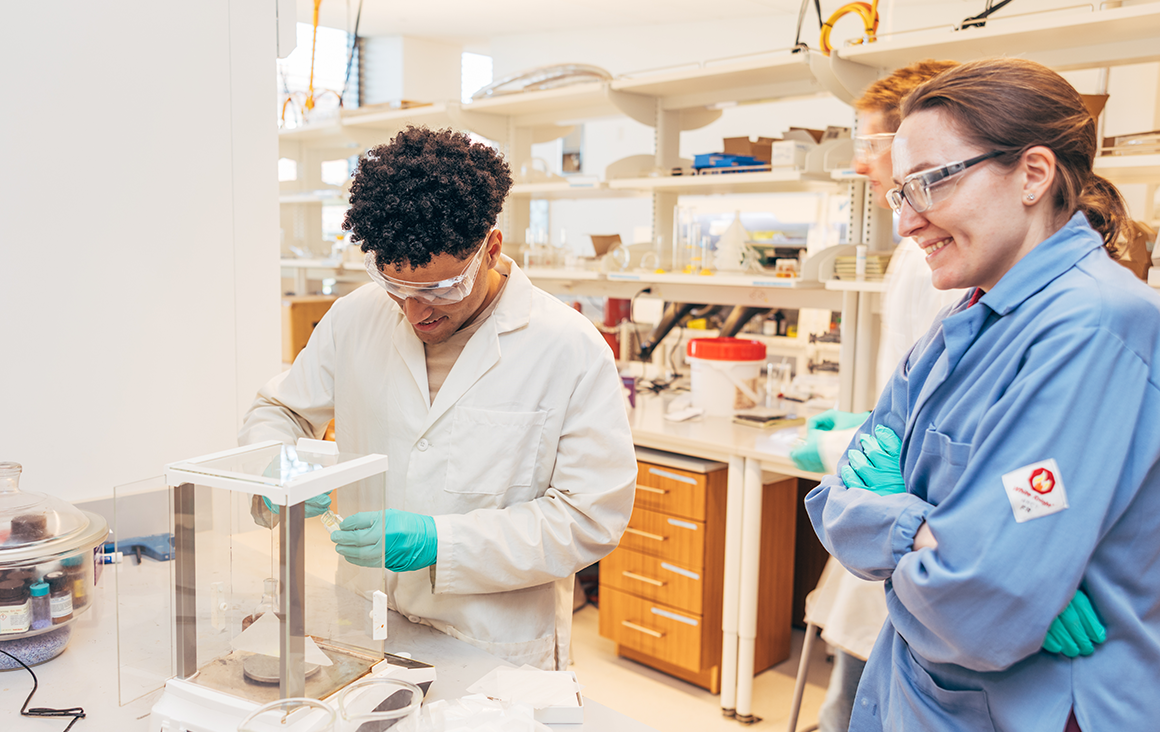 A female professor in a blue lab coat watches approvingly as a male student scoops a material out of a flask to weigh.