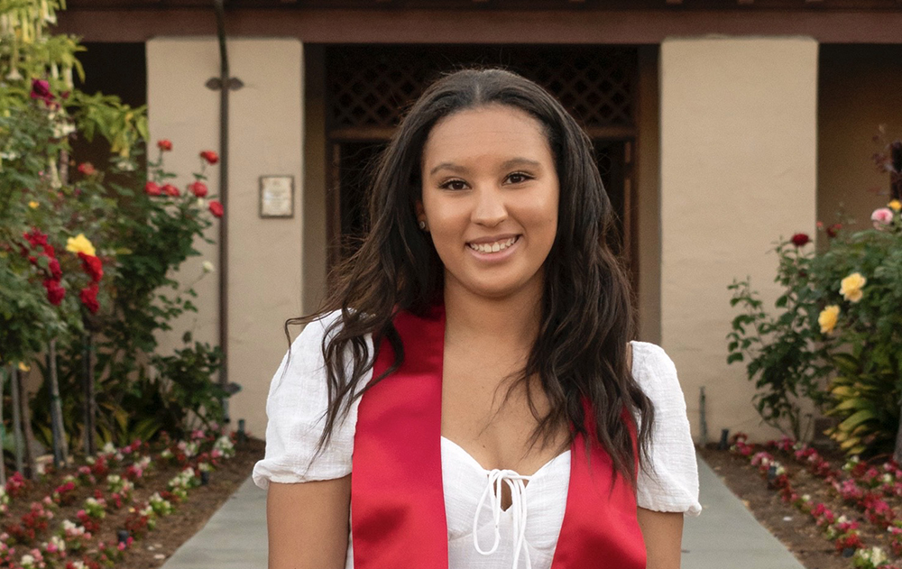 A person wearing a red stole and white top, smiling outdoors.