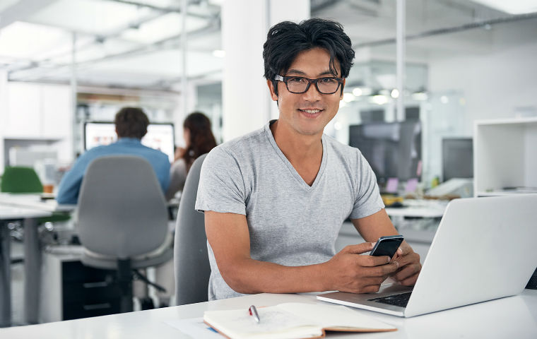 Young man with dark brown hair and glasses smiling from his desk. image link to story