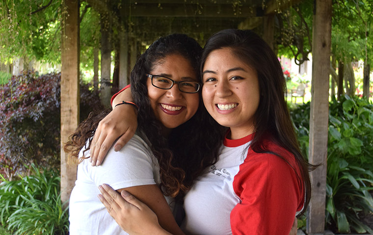Two people smiling and hugging under a green leafy canopy.