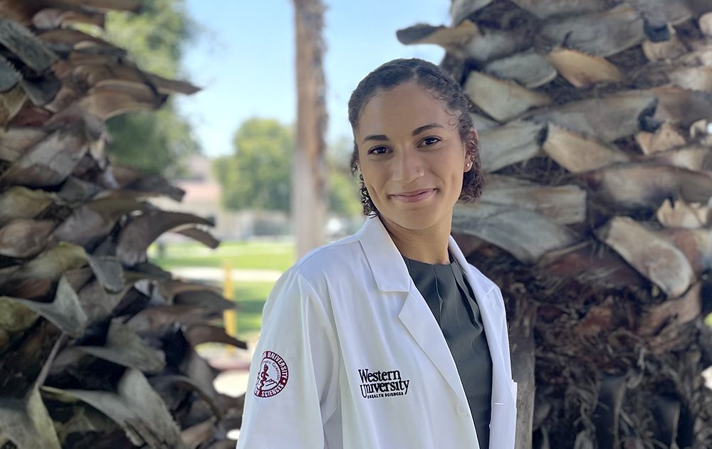 Noel Simms stands in her lab coat in front of palm trees on campus.