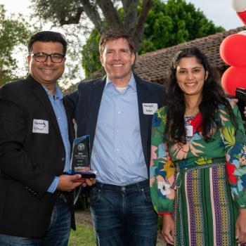 Three people smiling outdoors at an event with red and white balloons.
