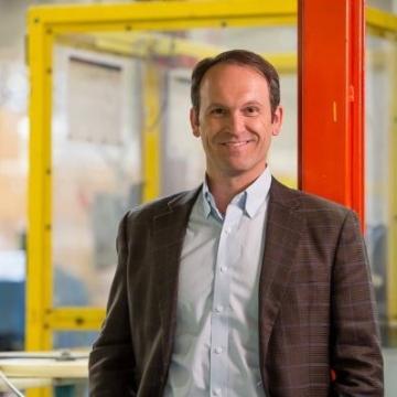 A man in a suit jacket standing in front of an industrial background.