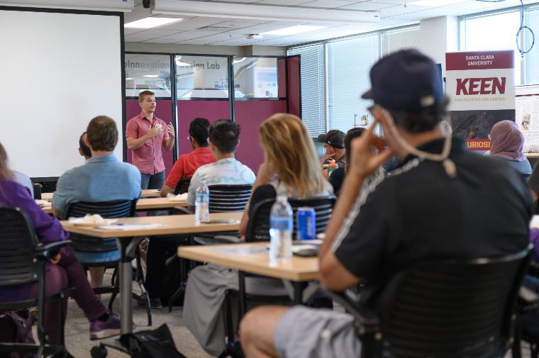 Alt text: Classroom setting with an instructor speaking to students at desks.