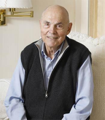 A man in a black vest with a striped shirt, seated indoors.