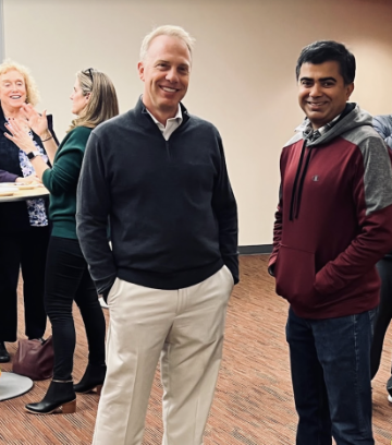 Two people standing indoors with a group in the background.