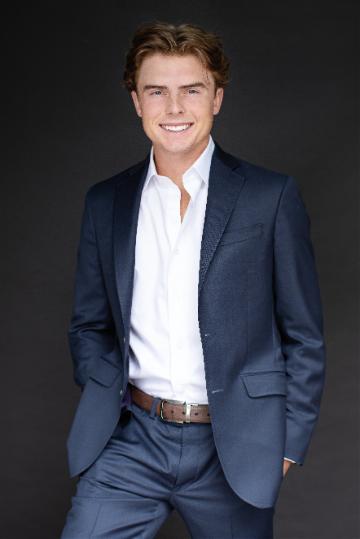 A person in a blue suit, smiling, against a dark background.