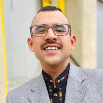 A person wearing glasses and a gray suit, standing outside near yellow flowers.