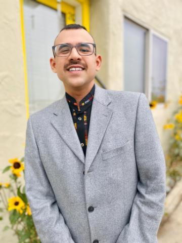 A person wearing glasses and a gray suit, standing outside near yellow flowers.